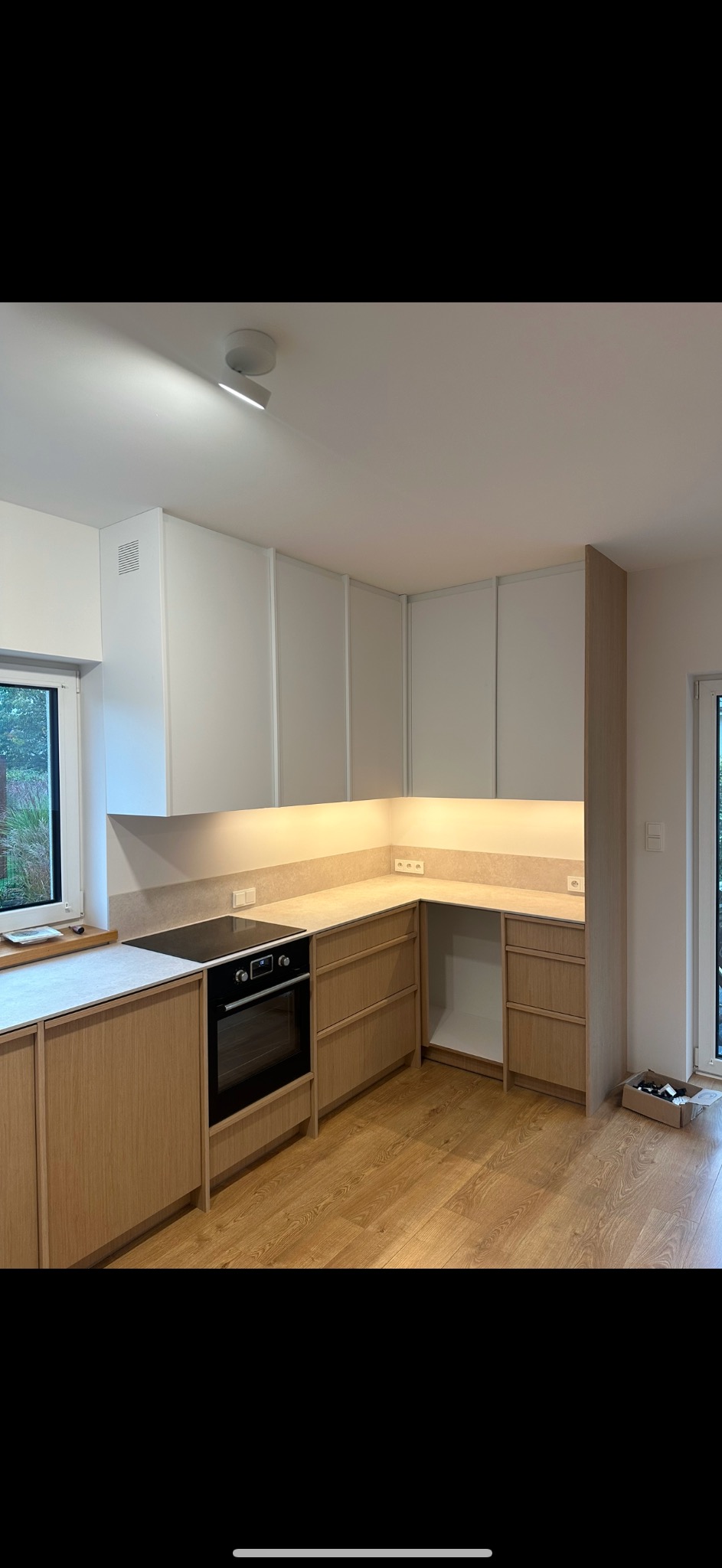 Jasna kuchnia z drewnianymi frontami szafek i białymi szafkami wiszącymi, oświetlona listwą LED pod szafkami, z płytą indukcyjną i piekarnikiem w zabudowie. Na podłodze panele imitujące drewno,...