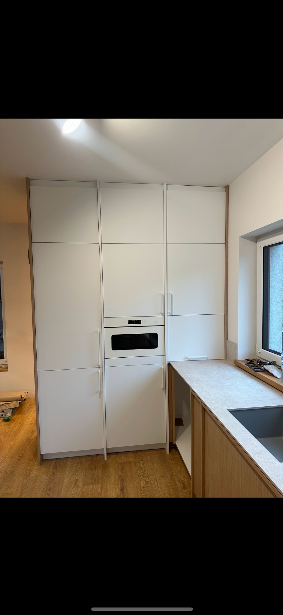 Nowoczesna zabudowa kuchenna z białymi frontami i drewnianymi akcentami, obejmująca szafki, piekarnik i blat z szarym zlewem, widoczna na tle parkietu.