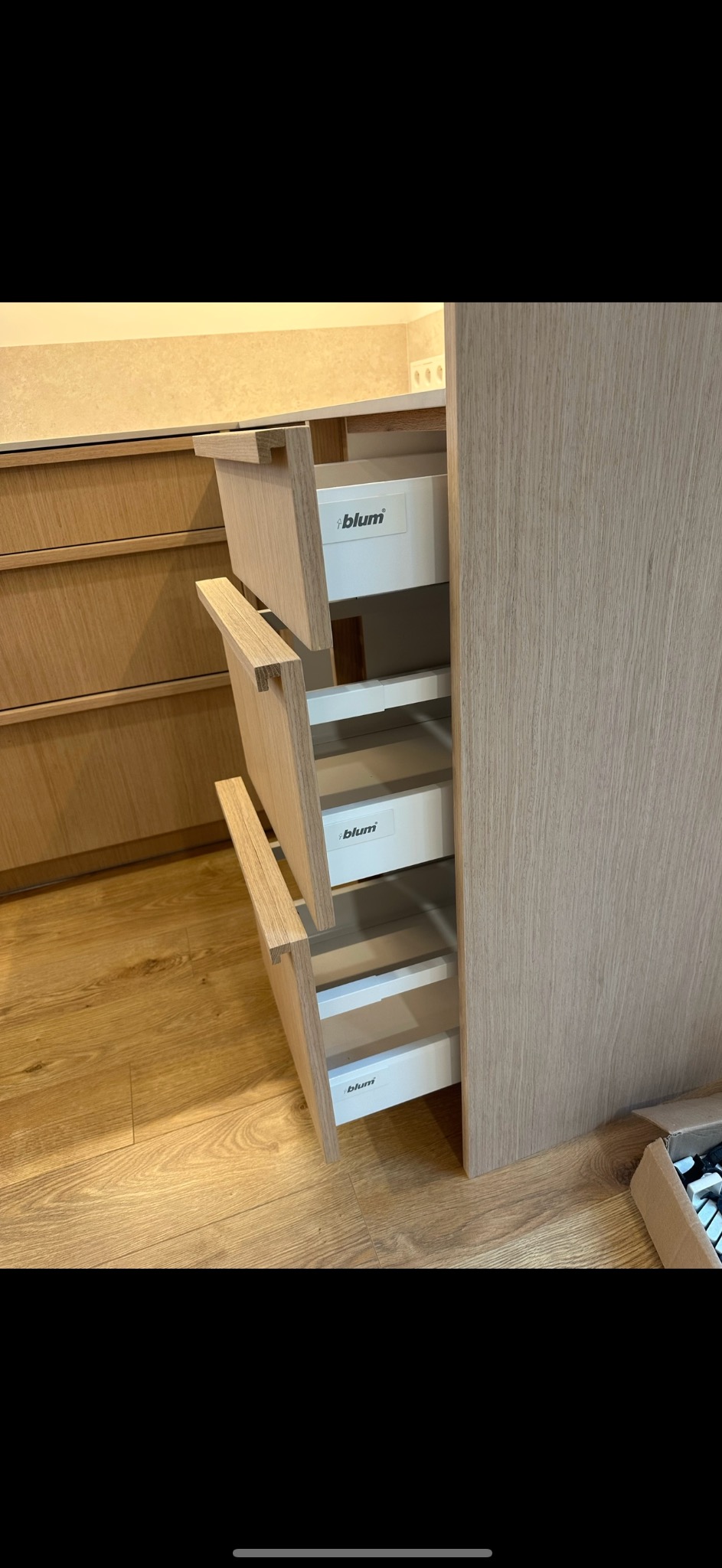 Jasna, drewniana szafka kuchenna z trzema wysuniętymi szufladami z systemem Blum, widoczne wnętrze szuflad i fragment podłogi oraz opakowanie akcesoriów.