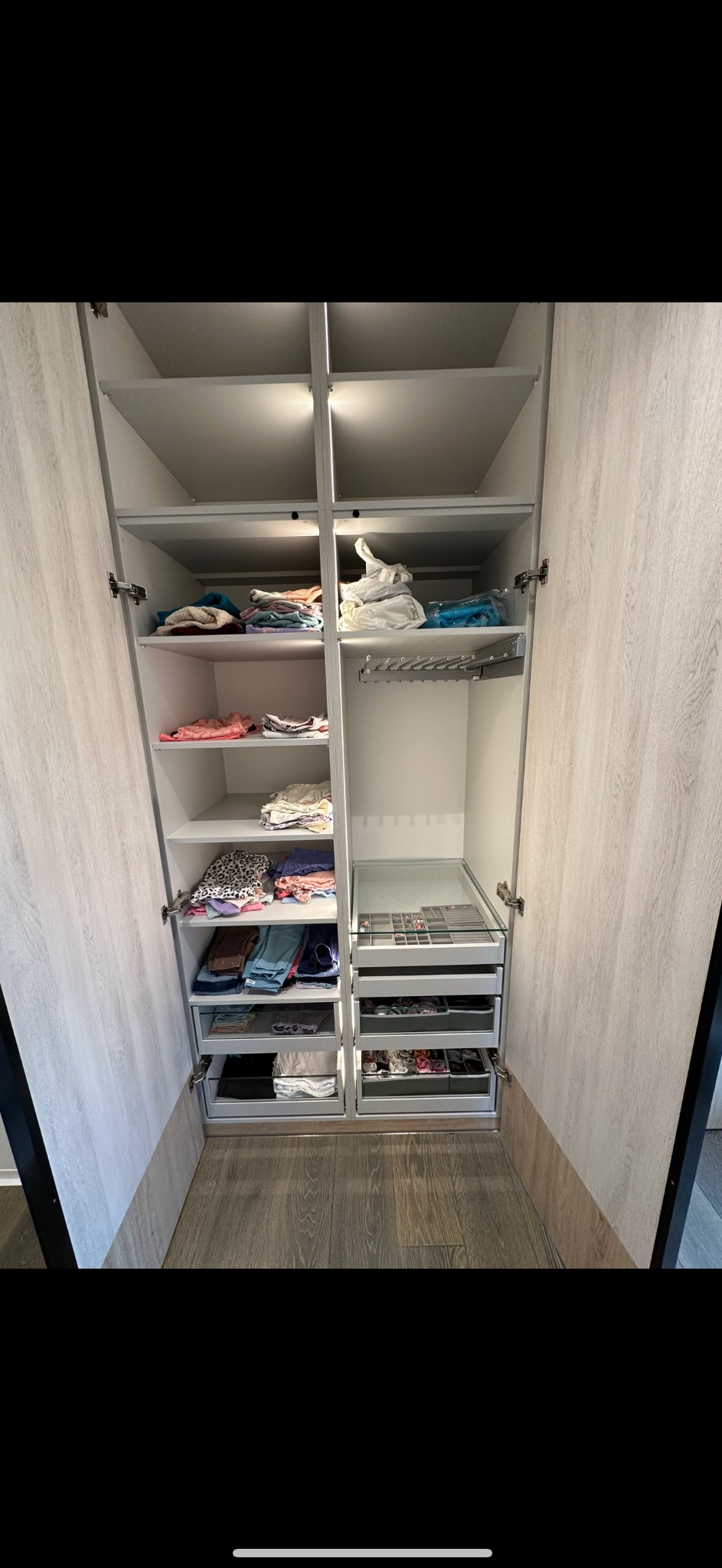 Otwarta szafa wnękowa z oświetleniem wewnętrznym, wyposażona w półki, drążki na ubrania i wysuwane szuflady z przegródkami na biżuterię i akcesoria; fronty szafy w kolorze jasnego drewna.