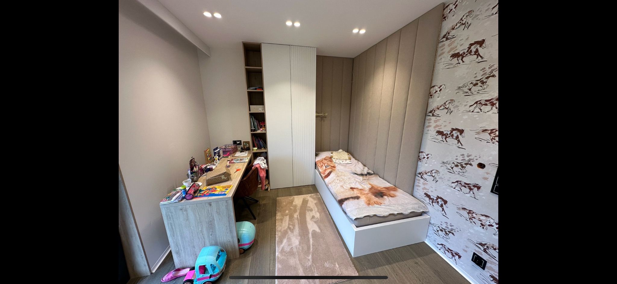 Pokój dziecięcy z zabudowaną szafą, biurkiem i łóżkiem z tapicerowanym zagłówkiem, tapeta w konie, zabawki rozrzucone na biurku i podłodze.