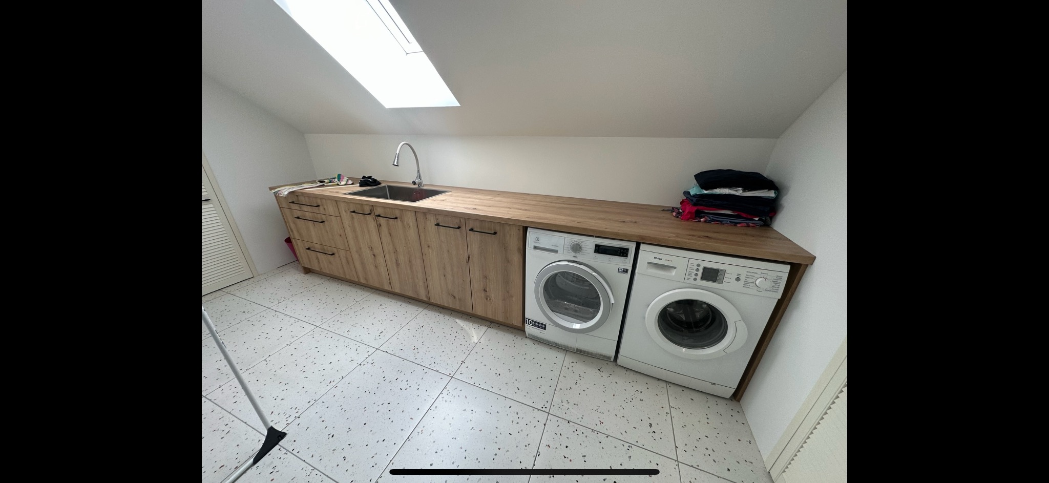 Pralka i suszarka zabudowane pod blatem z drewnianym wykończeniem, zintegrowane z szafkami i zlewem w pomieszczeniu gospodarczym na poddaszu ze świetlikiem.