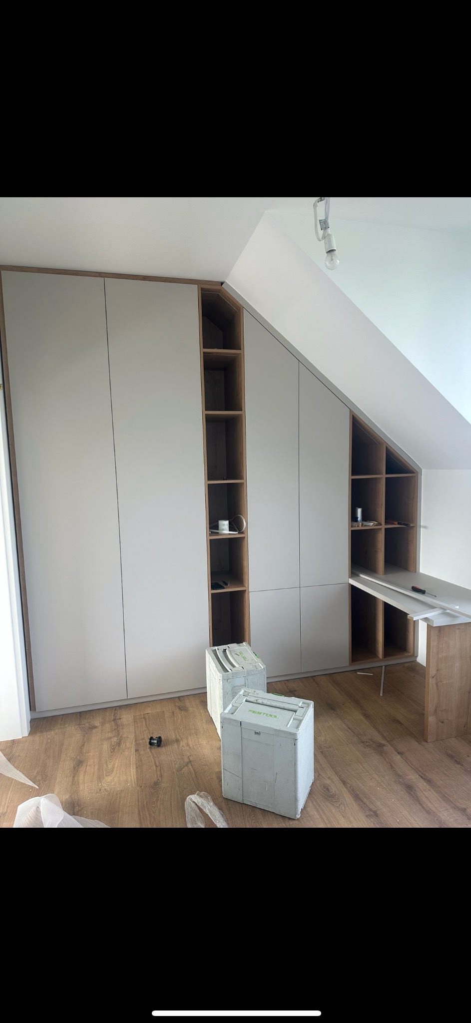 Montaż zabudowy wnękowej na poddaszu: szare fronty szaf z drewnianym obramowaniem, otwarte półki z drobnymi przedmiotami, skrzynki narzędziowe na podłodze.