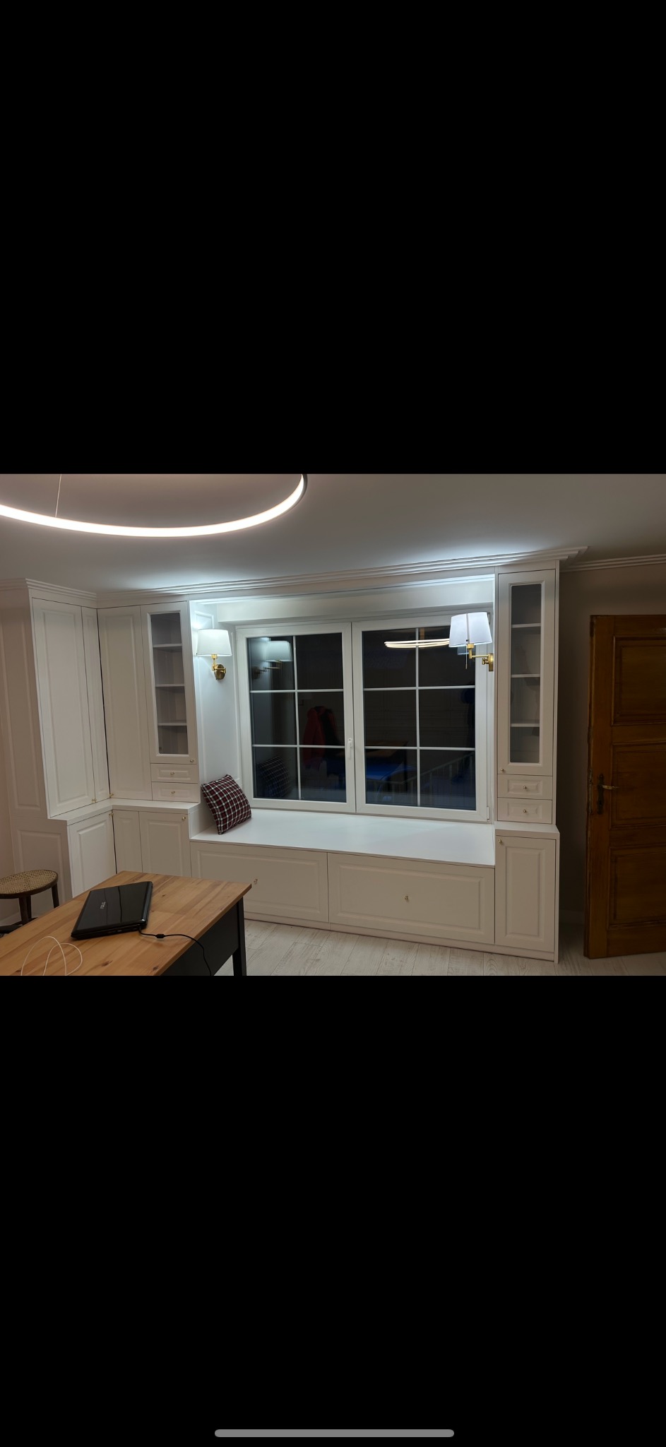 Biała zabudowa okienna z szafkami i siedziskiem pod oknem, uzupełniona o szafy po bokach, oświetlona złotymi kinkietami, w tle widoczne okno z widokiem na zewnątrz.