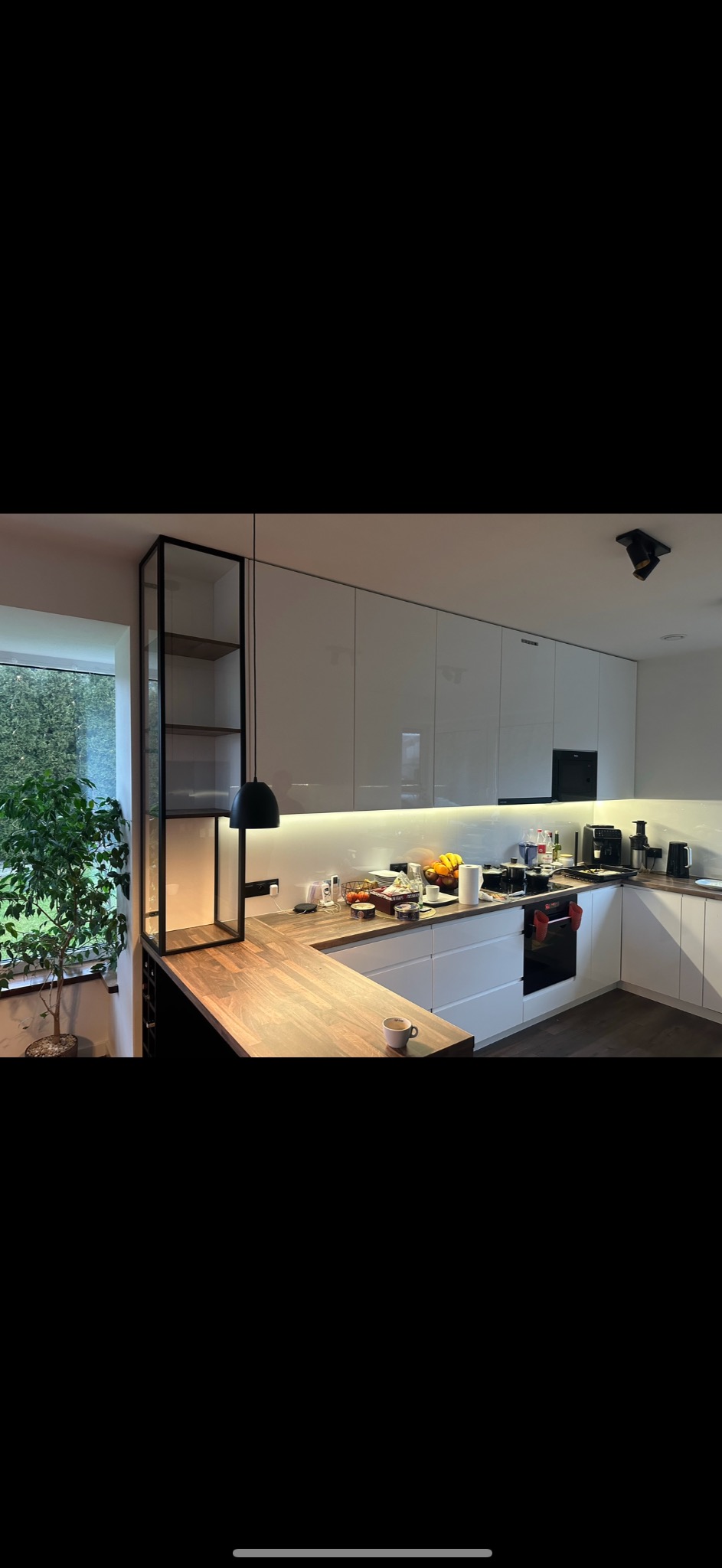 Nowoczesna kuchnia z białymi, lakierowanymi frontami szafek, drewnianym blatem i czarną, minimalistyczną lampą wiszącą, z widocznym fragmentem przeszklonej witryny i rośliną doniczkową przy oknie.