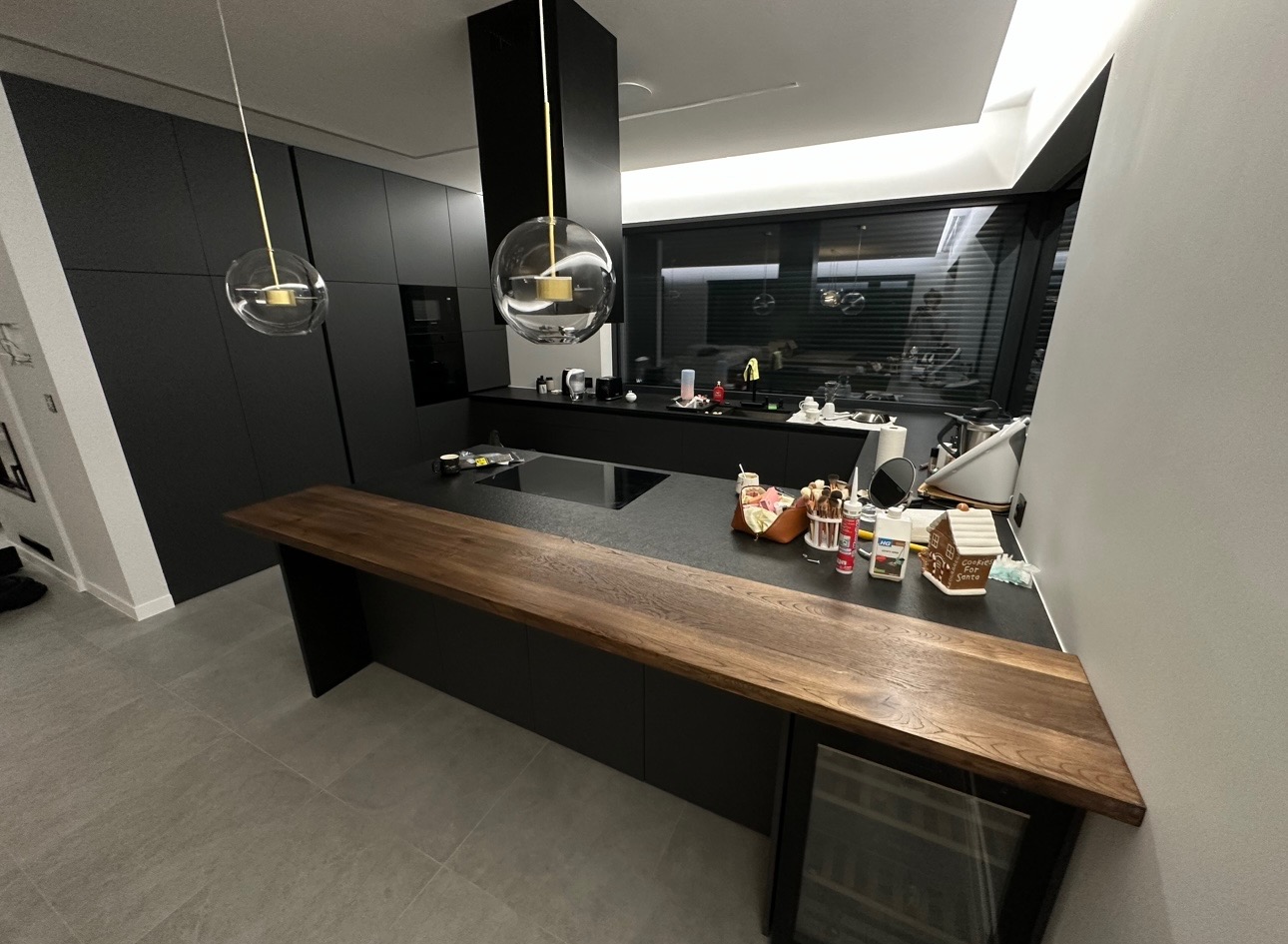 Nowoczesna czarna kuchnia z drewnianym blatem barowym, oświetlona wiszącymi lampami kulami, widok na okno i nieporządek na blacie.