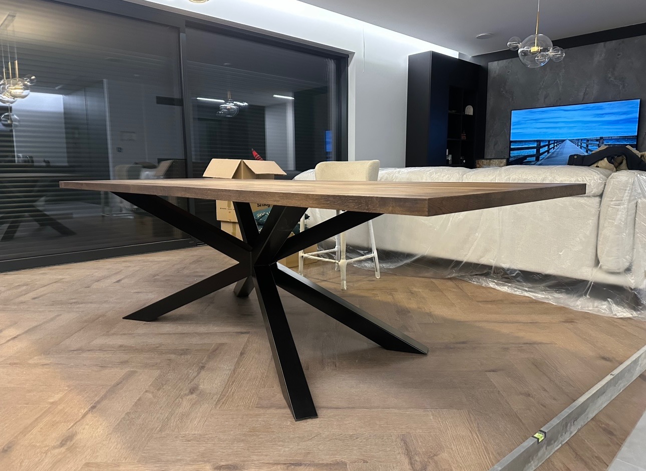 Stół z blatem z ciemnego drewna i czarną, geometryczną podstawą w nowoczesnym wnętrzu z dużym oknem przesuwnym i drewnianą podłogą w jodełkę. W tle widoczna sofa zabezpieczona folią malarską.