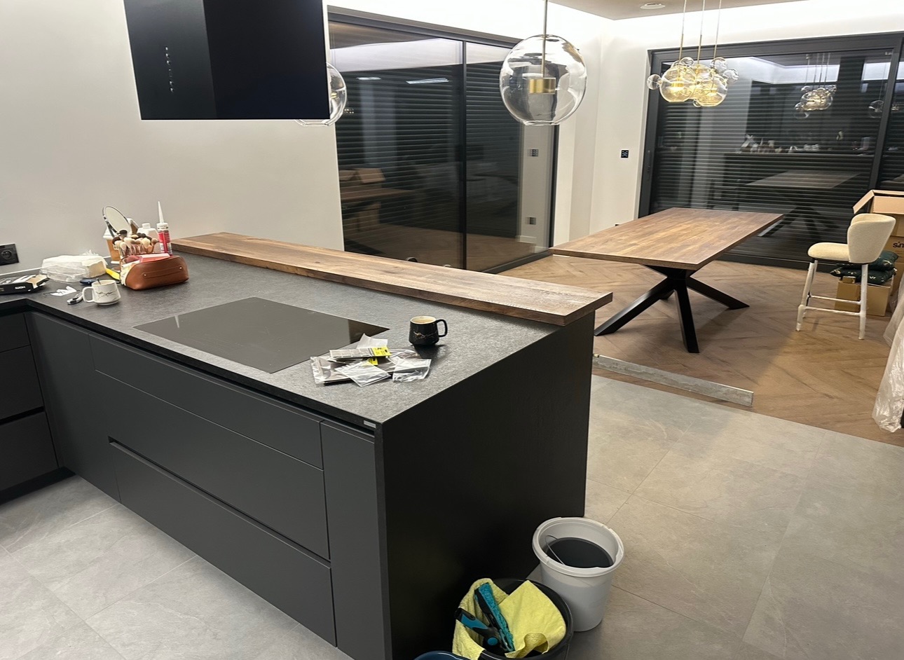 Częściowy widok nowoczesnej kuchni z ciemnymi szafkami i blatem, na którym leżą akcesoria montażowe i stoi kubek, w tle stół z drewnianym blatem na metalowej podstawie i krzesło barowe, widoczne...