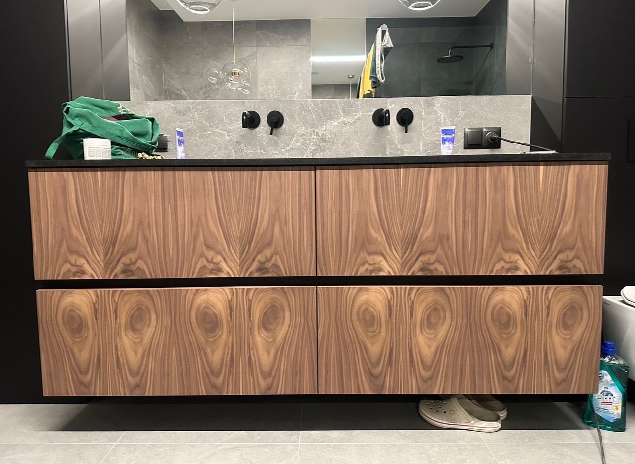 Wnętrze łazienki z szafką pod umywalką z forniru orzechowego, czarny blat, czarne baterie umywalkowe, szare płytki na ścianie, widoczne akcesoria łazienkowe i obuwie pod szafką.