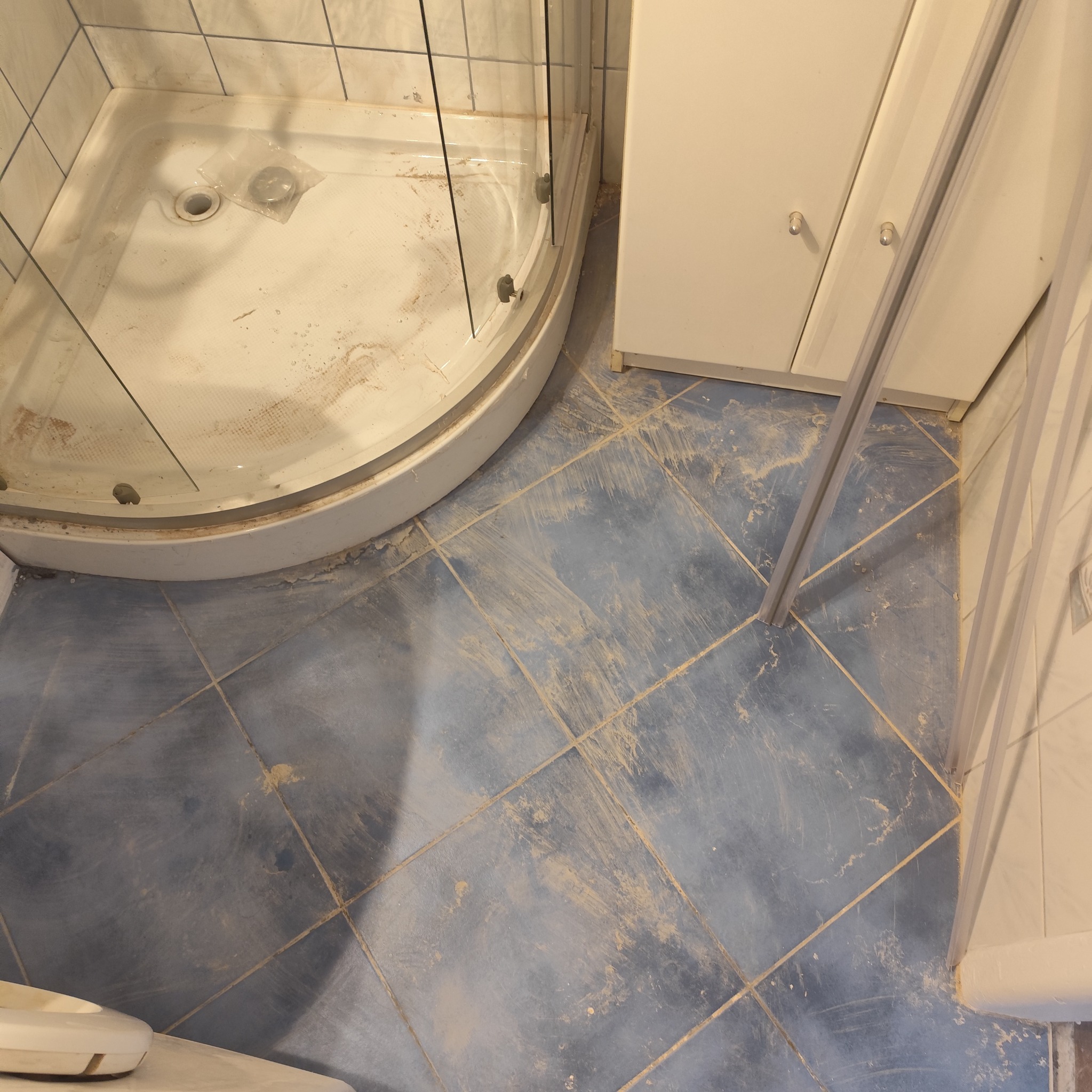 Zabrudzona kabina prysznicowa z niebieskimi płytkami podłogowymi po remoncie. Widoczne pozostałości po pracach budowlanych. Ujęcie z góry, brak osób.