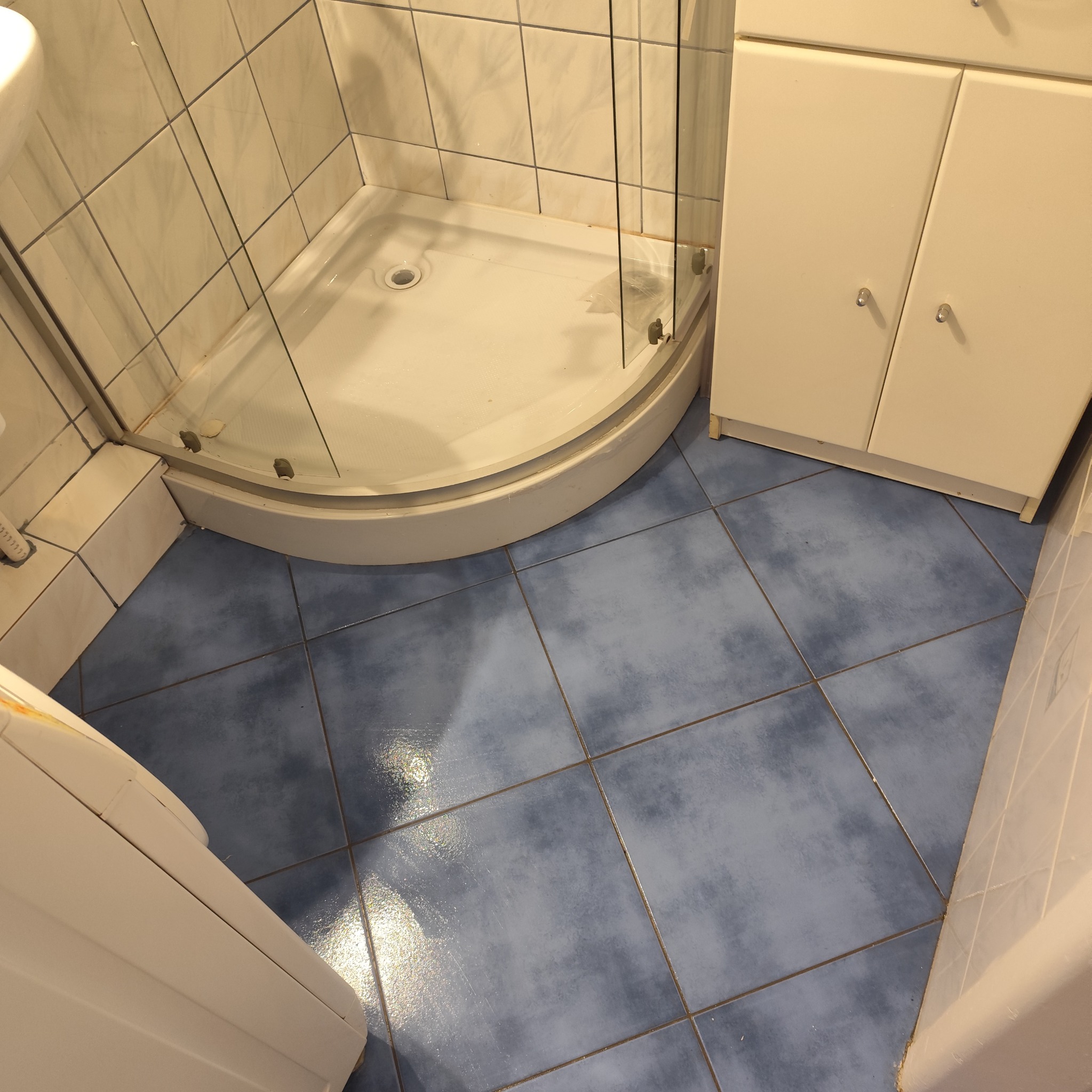 Umyta kabina prysznicowa z niebieską podłogą z płytek i szafką w łazience. Widoczny połysk na płytkach po sprzątaniu, czysta powierzchnia.