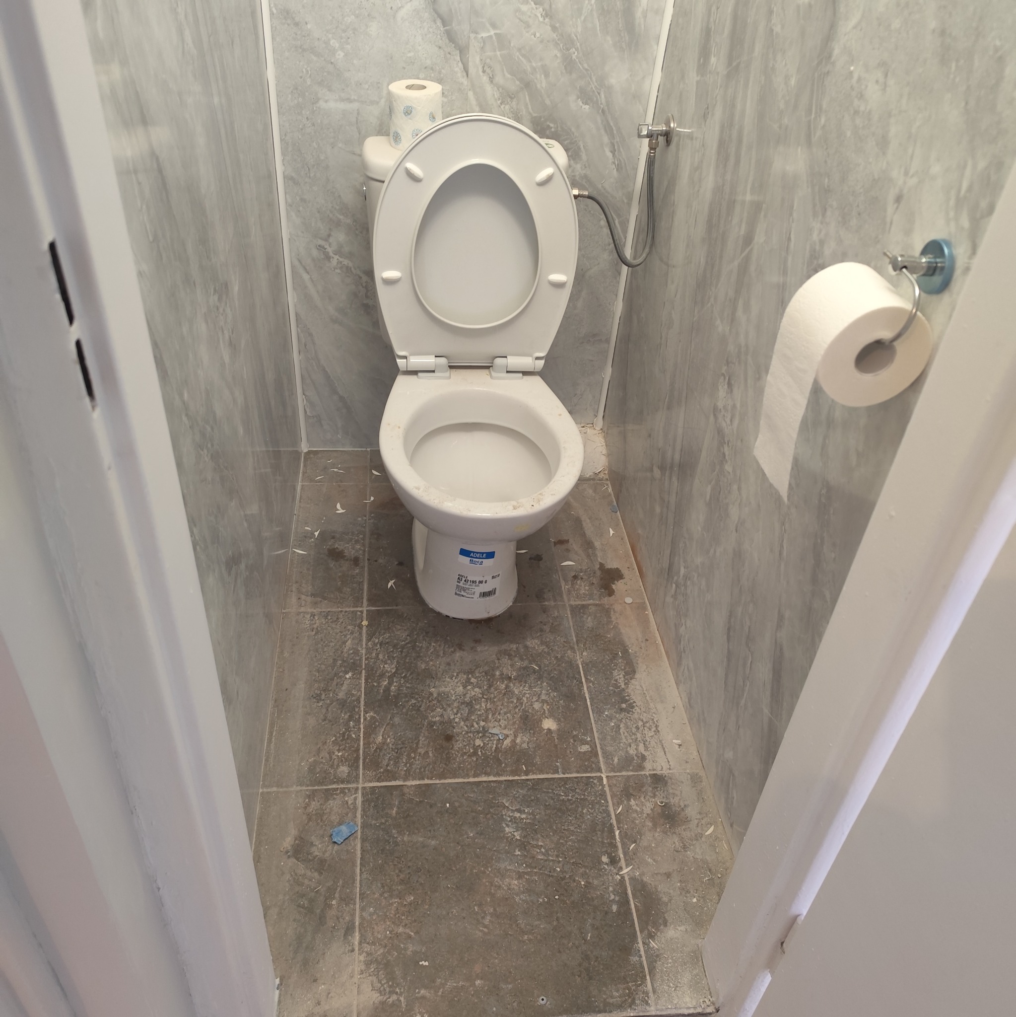 Toaleta z otwartą klapą w wąskim pomieszczeniu wyłożonym szarymi płytkami imitującymi marmur, widoczne zabrudzenia na podłodze i ścianach. Ujęcie zza futryny drzwi.