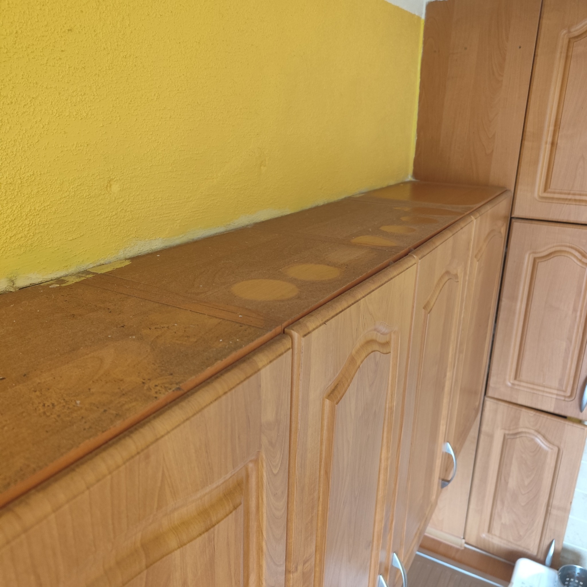 Zakurzone, drewniane szafki kuchenne pod żółtą ścianą. Na blacie widoczne okrągłe ślady po naczyniach, sugerujące potrzebę odkurzenia i umycia.