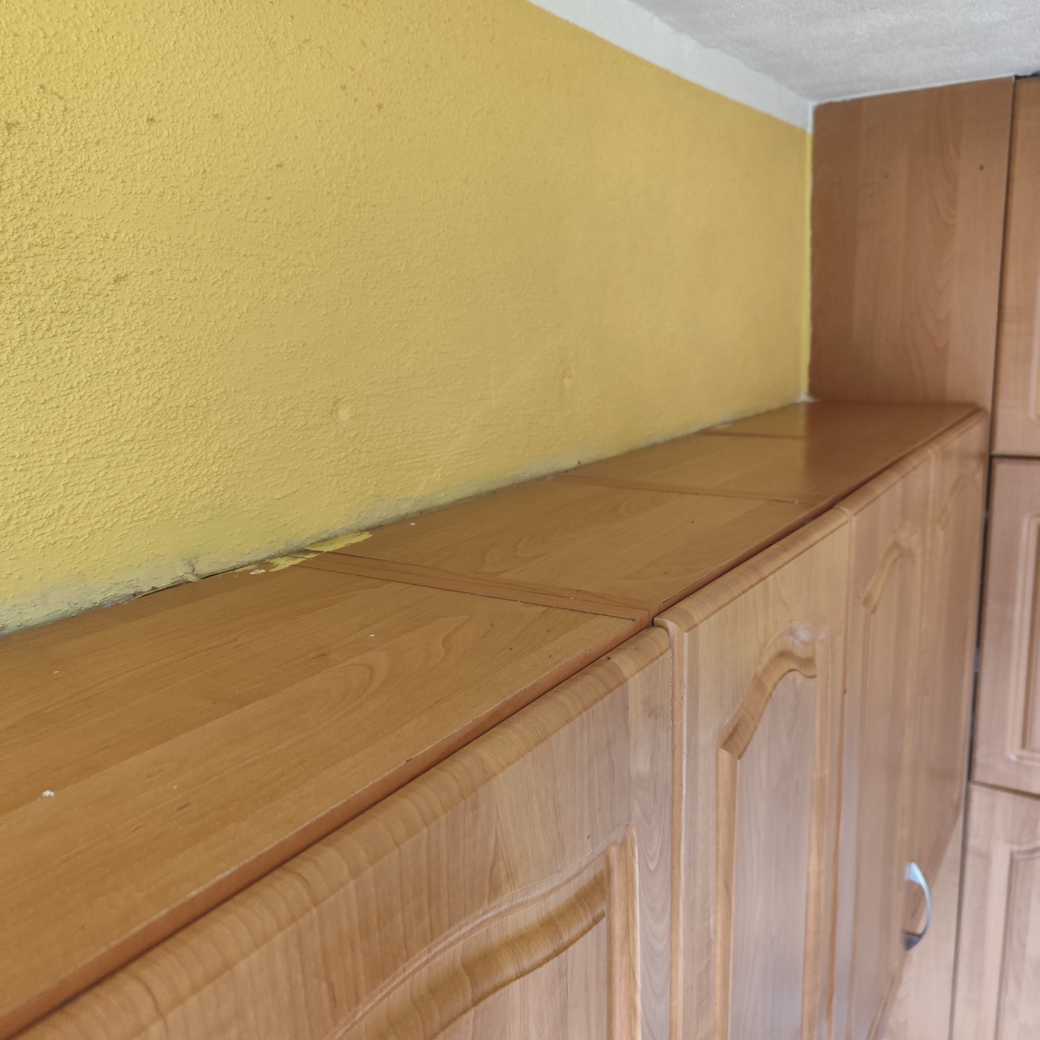 Fragment szafki wiszącej z drewnianymi frontami i żółtą ścianą o nierównej fakturze w tle. Widoczne drobne zabrudzenia na szafce i ślady malowania na styku ściany i szafki.