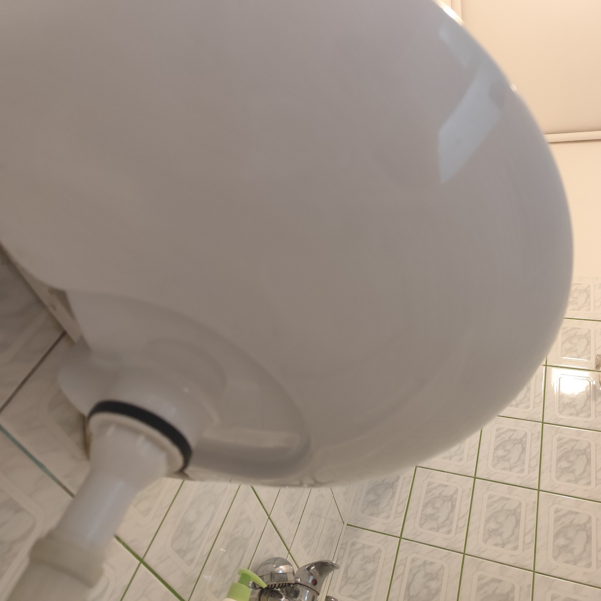 Biała umywalka z widocznym syfonem i baterią, na tle płytek ściennych. Ujęcie z dołu, podkreślające detale instalacji. Widoczny dozownik mydła.