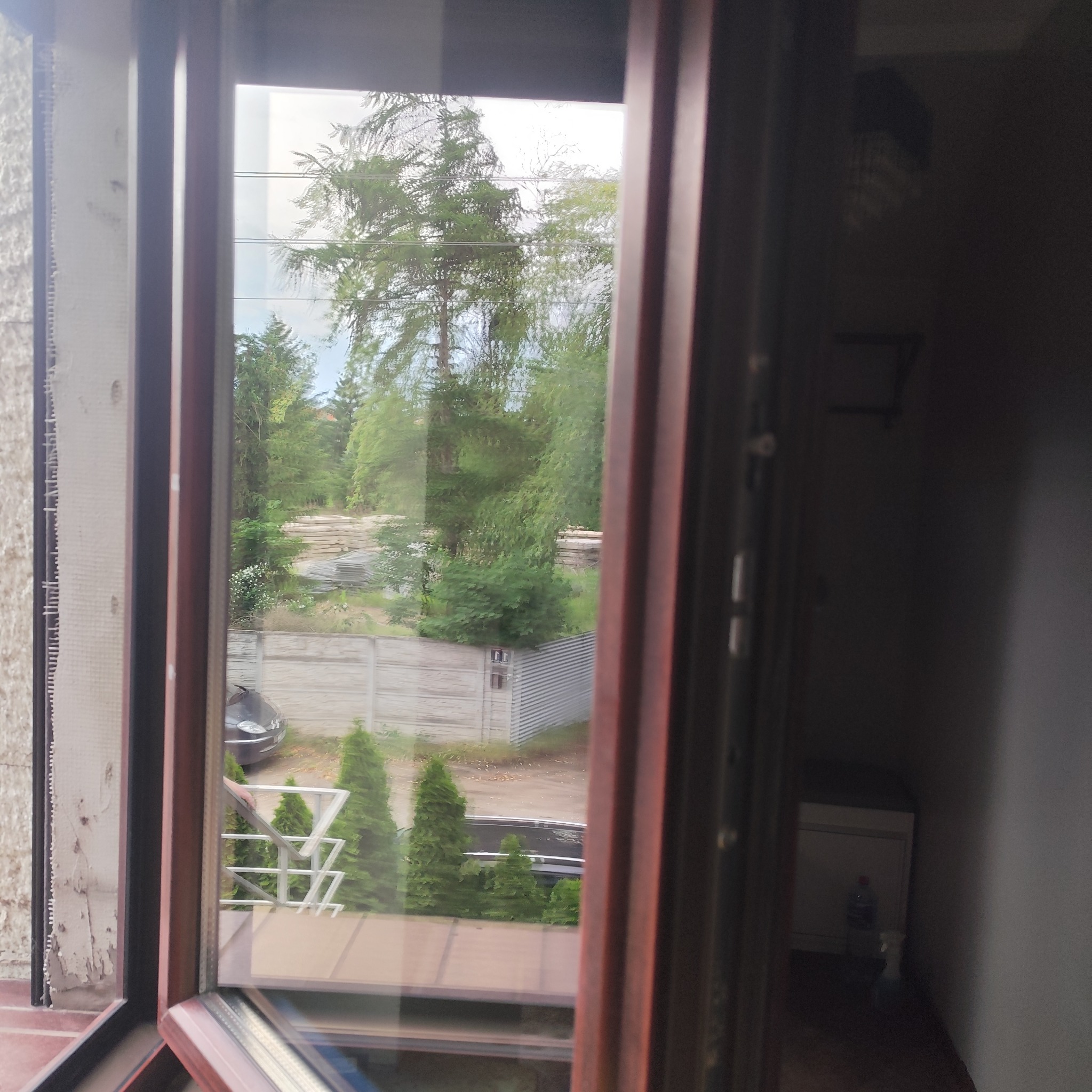 Otwarty widok z okna na zewnątrz, z fragmentem ściany po lewej i korytarzem po prawej. Widoczny fragment balkonu i zieleni za oknem.