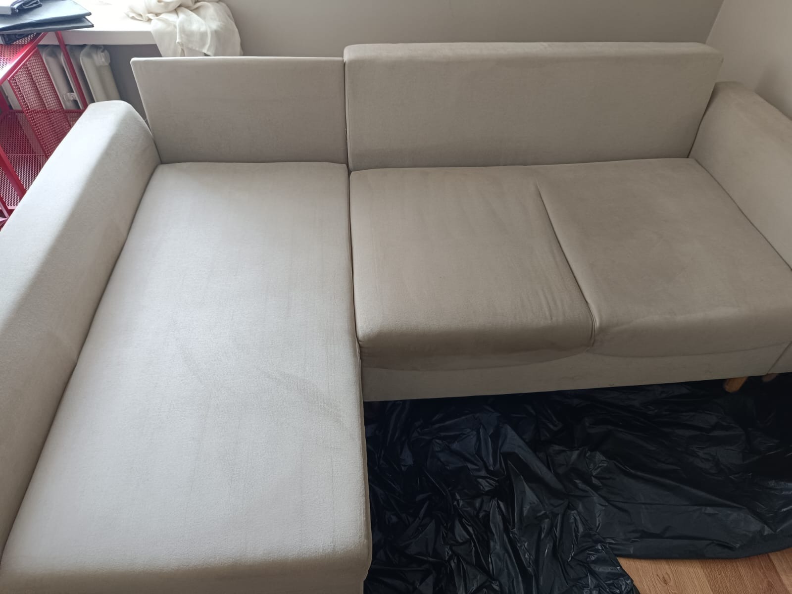 Jasnobeżowa sofa narożna po praniu tapicerki. Widoczna wyraźna różnica w czystości między wypraną a niewypraną częścią. Zabezpieczenie podłogi czarną folią.