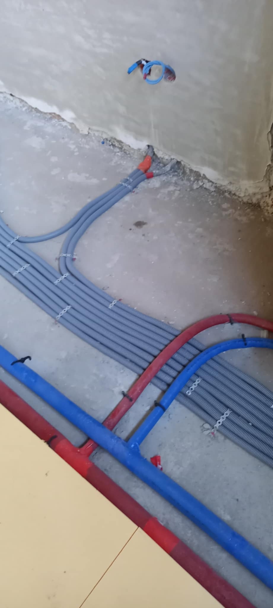Szare rury karbowane i niebiesko-czerwone rury wodne ułożone na podłodze w surowym stanie, przymocowane do podłoża. Widoczne fragmenty ścian w tle.