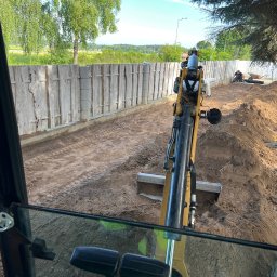 4HOME - Widok z kabiny koparki na wykop wzdłuż ogrodzenia. W tle pracownicy i narzędzia. Świeży wykop z widocznymi śladami pracy maszyny.