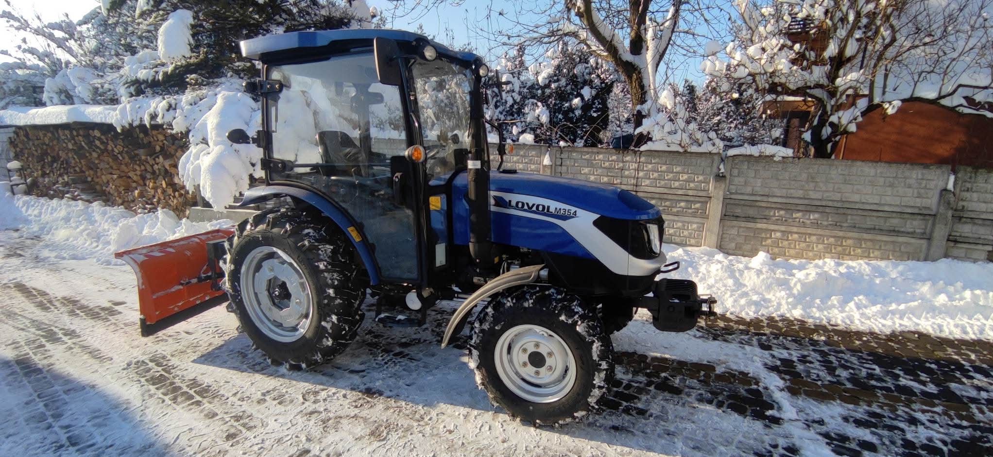 Niebieski traktor Lovol M354 z pługiem do śniegu na ośnieżonej drodze, w tle drewno i betonowe ogrodzenie. Słoneczny zimowy dzień.