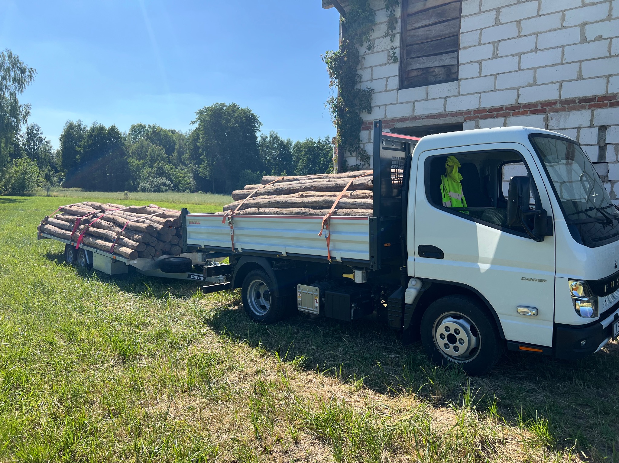 Ciężarówka z przyczepą wypełniona palami drewnianymi na tle budynku z cegły i zieleni, w słoneczny dzień. Transport drewna.
