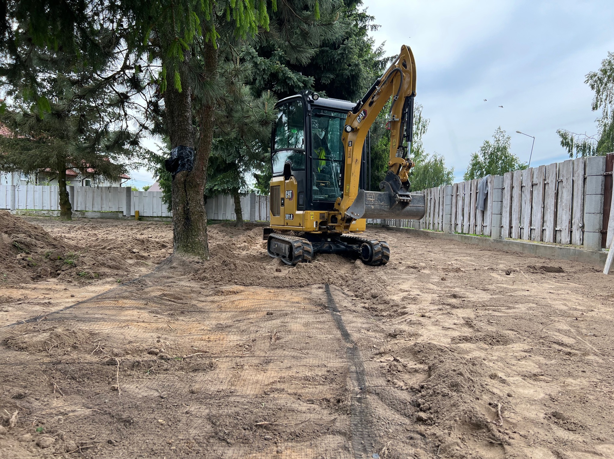 Żółta mini koparka CAT 301.8 na placu budowy z drzewami i betonowym ogrodzeniem w tle. Widoczne ślady pracy i siatka wzmacniająca grunt.