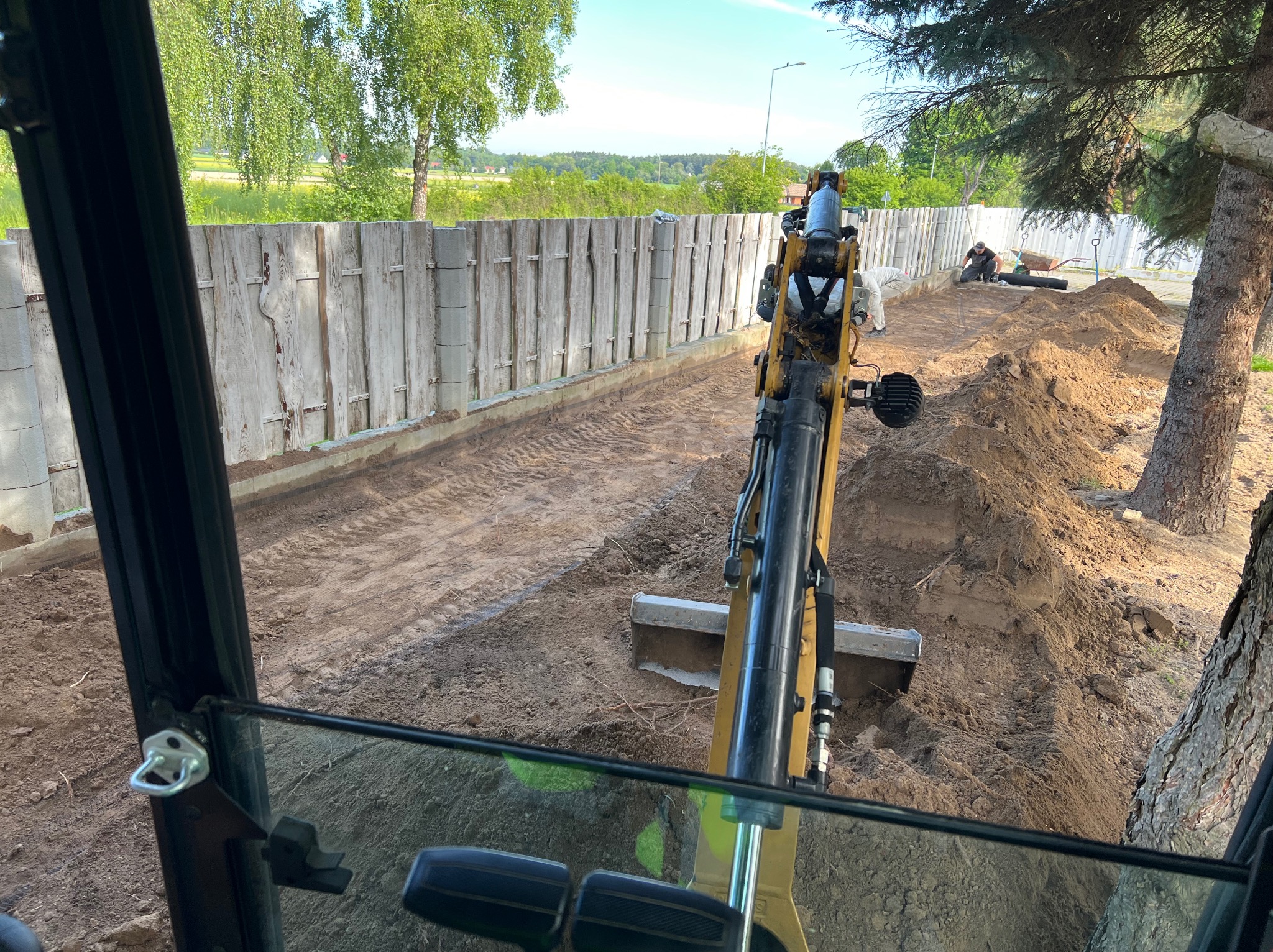 Widok z kabiny koparki na wykop wzdłuż ogrodzenia. W tle pracownicy i narzędzia. Świeży wykop z widocznymi śladami pracy maszyny.
