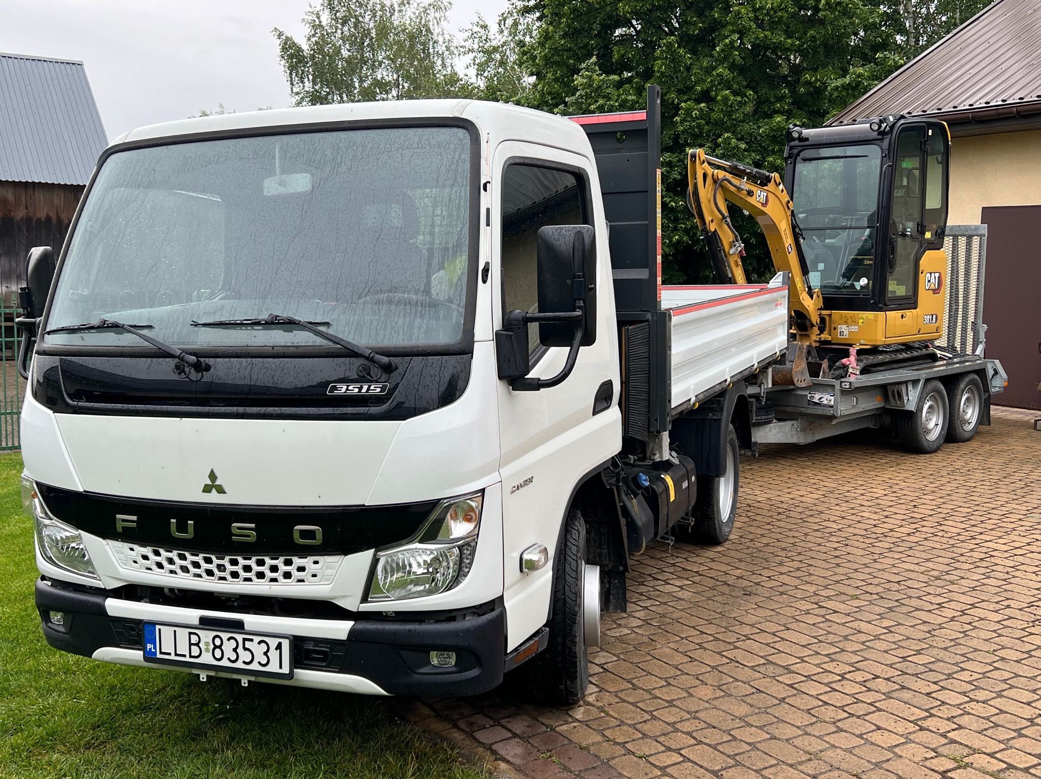 Biały samochód ciężarowy Fuso z żółtą mini koparką CAT 301.8 na przyczepie, zaparkowany na brukowanej nawierzchni. Widoczne drzewa i budynek w tle.
