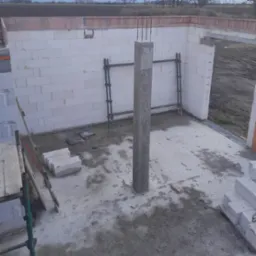 Wnętrze w trakcie budowy: ściany z białych bloczków, betonowy słup wzmocniony stalą, widoczne rusztowania. Surowy stan, przygotowanie do dalszych prac.