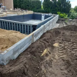 Budowa basenu: betonowa konstrukcja z szalunkiem, obok ziemia i piasek. Widoczne etapy prac ziemnych i murowania. Krajobraz z zielenią w tle.