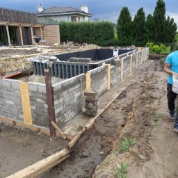 Usługi Ogólnobudowlane - Budowa fundamentów basenu z szarej cegły, widoczne szalunki i zbrojenie. W tle dom i zieleń. Etap prac murarskich, przygotowanie do wylania betonu.