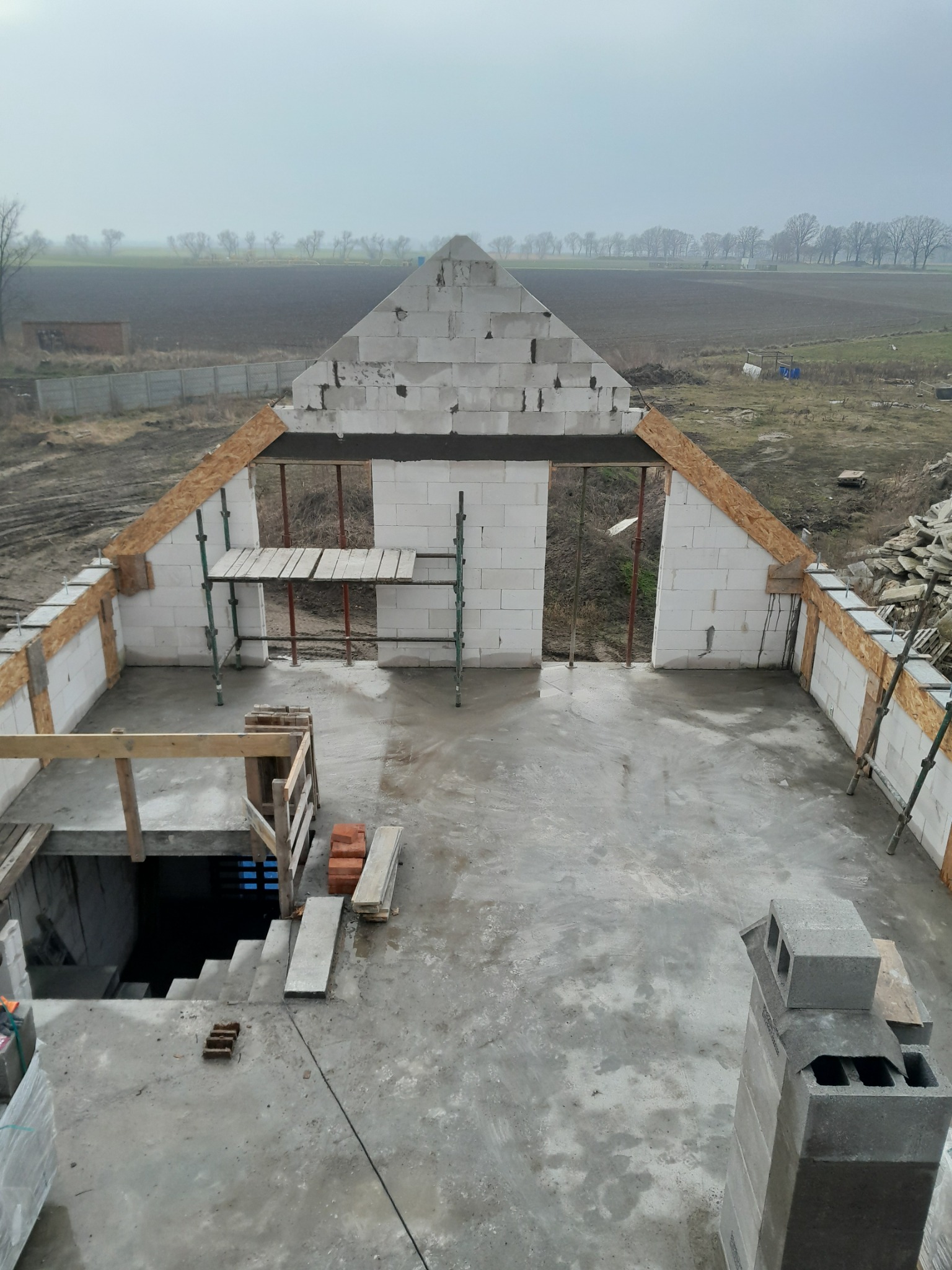 Wysoki kąt na dom w trakcie budowy z betonu komórkowego. Widoczne rusztowania, otwory okienne i wejście do piwnicy. Krajobraz w tle.