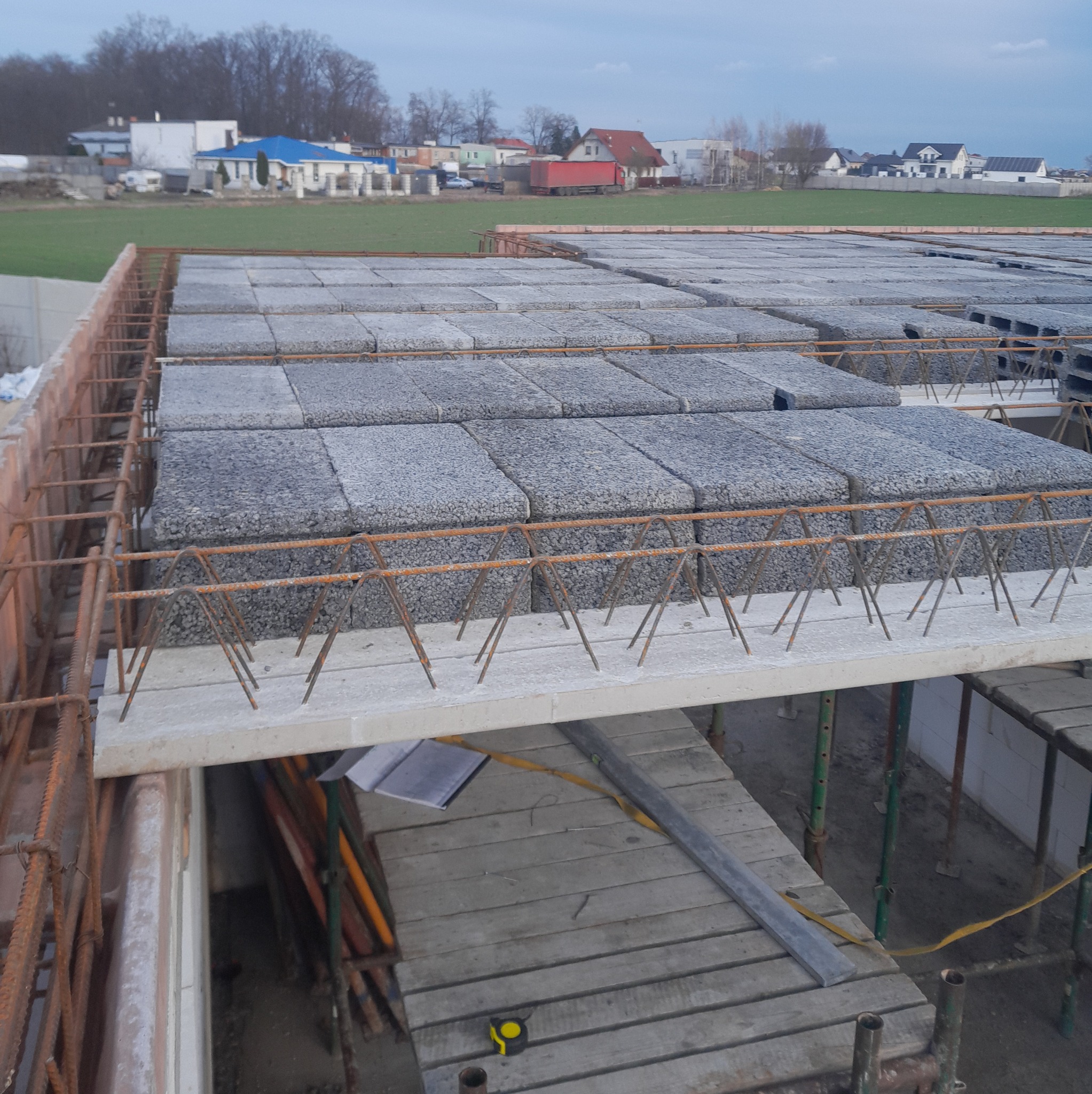 Montaż stropu Teriva z pustaków szalunkowych i zbrojenia na budowie domu, widok z góry na przygotowany strop przed zalaniem betonem. W tle zabudowa mieszkalna.