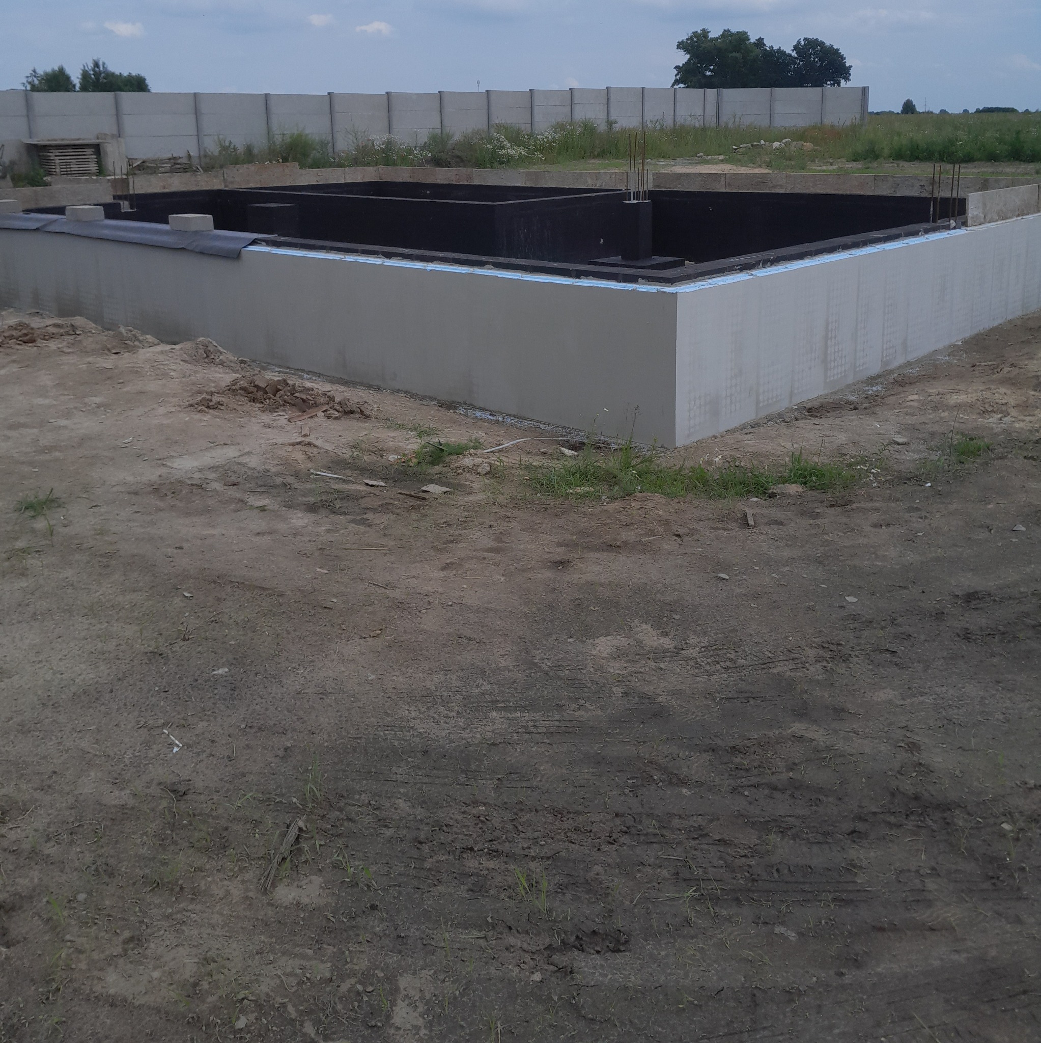 Zabezpieczony fundament domu z izolacją termiczną i przeciwwodną, przygotowany do dalszych prac budowlanych. Widoczne zbrojenia i teren budowy.