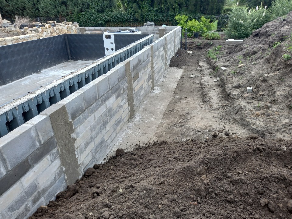 Budowa basenu: Ściany z szarej cegły, zbrojenie wewnątrz. Otoczenie to ziemia i zieleń. Widoczne etapy prac murarskich i przygotowania do wylania betonu.