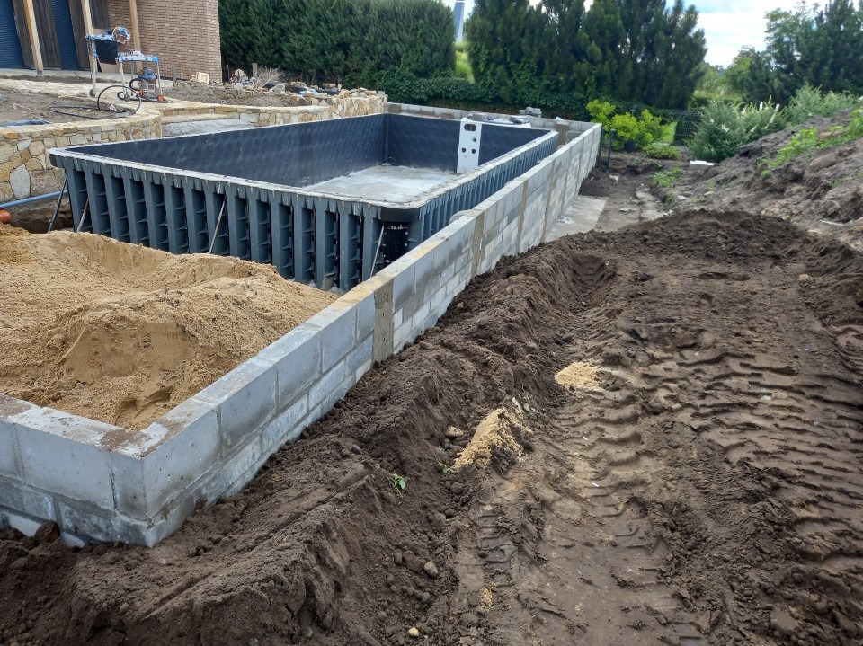 Budowa basenu: betonowa konstrukcja z szalunkiem, obok ziemia i piasek. Widoczne etapy prac ziemnych i murowania. Krajobraz z zielenią w tle.