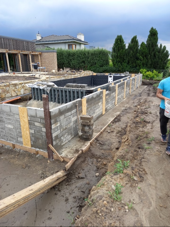 Budowa fundamentów basenu z szarej cegły, widoczne szalunki i zbrojenie. W tle dom i zieleń. Etap prac murarskich, przygotowanie do wylania betonu.
