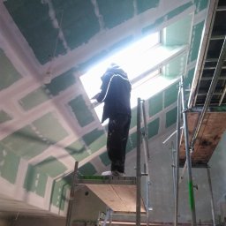 Paul Construction Paul Gharajlou - Pracownik na rusztowaniu szpachluje łączenia płyt gipsowo-kartonowych na skośnym suficie z oknem dachowym, widoczne ślady po szpachlowaniu.
