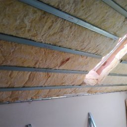 Paul Construction Paul Gharajlou - Izolacja poddasza wełną mineralną, widoczne metalowe profile montażowe i fragment ściany, etap prac wykończeniowych.