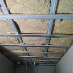 Sufit podwieszany w trakcie montażu: metalowa konstrukcja z widoczną izolacją z wełny mineralnej, przewody wentylacyjne wystające z sufitu.