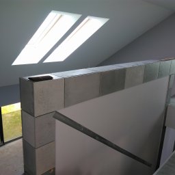 Paul Construction Paul Gharajlou - Wnętrze domu w trakcie wykańczania: betonowe bloczki tworzą ściankę działową pod skosem dachu z oknami, widoczne surowe ściany i podłoga w szarych płytkach, w tle fragment pomieszczenia w trakcie...