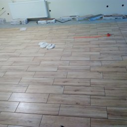 Paul Construction Paul Gharajlou - Ułożona podłoga z płytek imitujących drewno, widoczne pudełka po płytkach przy ścianie, pomarańczowa miarka i czerwona taśma malarska, oraz białe rękawice robocze.