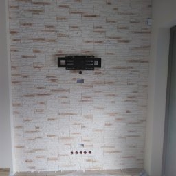 Paul Construction Paul Gharajlou - Ściana wyłożona dekoracyjną cegłą w odcieniach bieli i beżu, z zamontowanym uchwytem do telewizora i przygotowanymi otworami na przewody, zabezpieczona folią malarską podczas prac wykończeniowych.