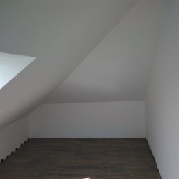 Paul Construction Paul Gharajlou - Poddasze po remoncie z nową podłogą z paneli o ciemnym odcieniu drewna i świeżo pomalowanymi na biało ścianami, widoczne listwy przypodłogowe w trakcie montażu.