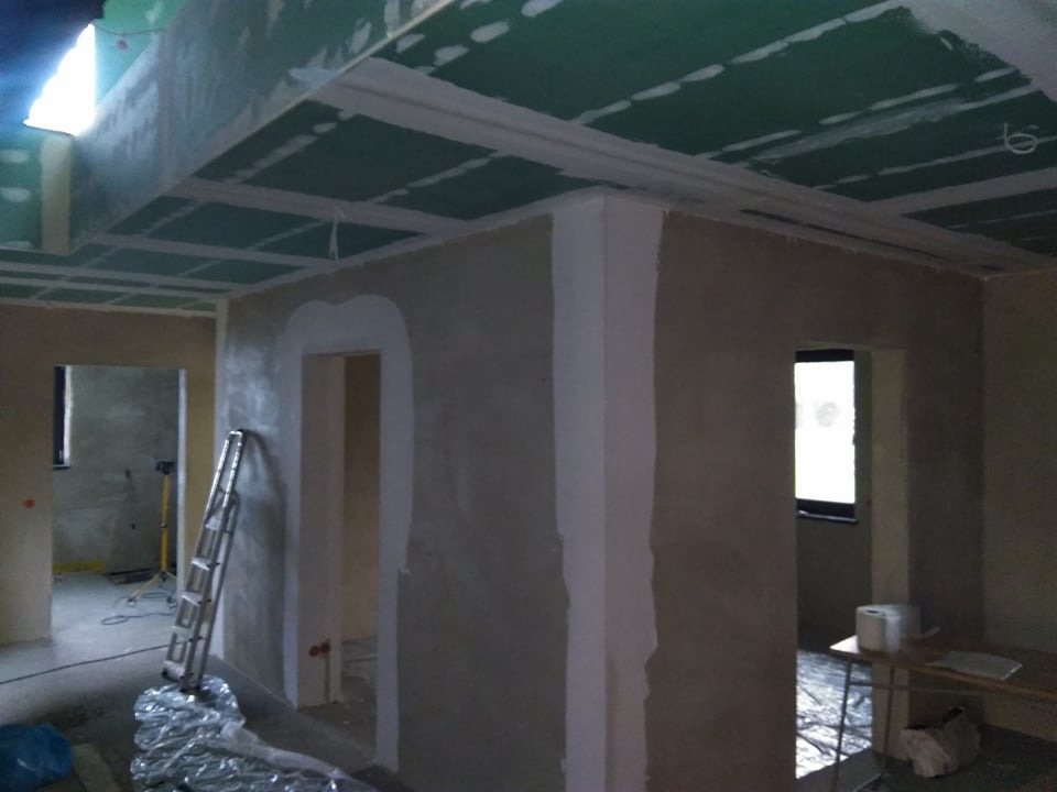 Wnętrze w trakcie prac wykończeniowych: szpachlowane ściany, zielone płyty gipsowo-kartonowe na suficie, aluminiowa drabina oparta o ścianę, widoczne profile sufitowe i przygotowane otwory drzwiowe.