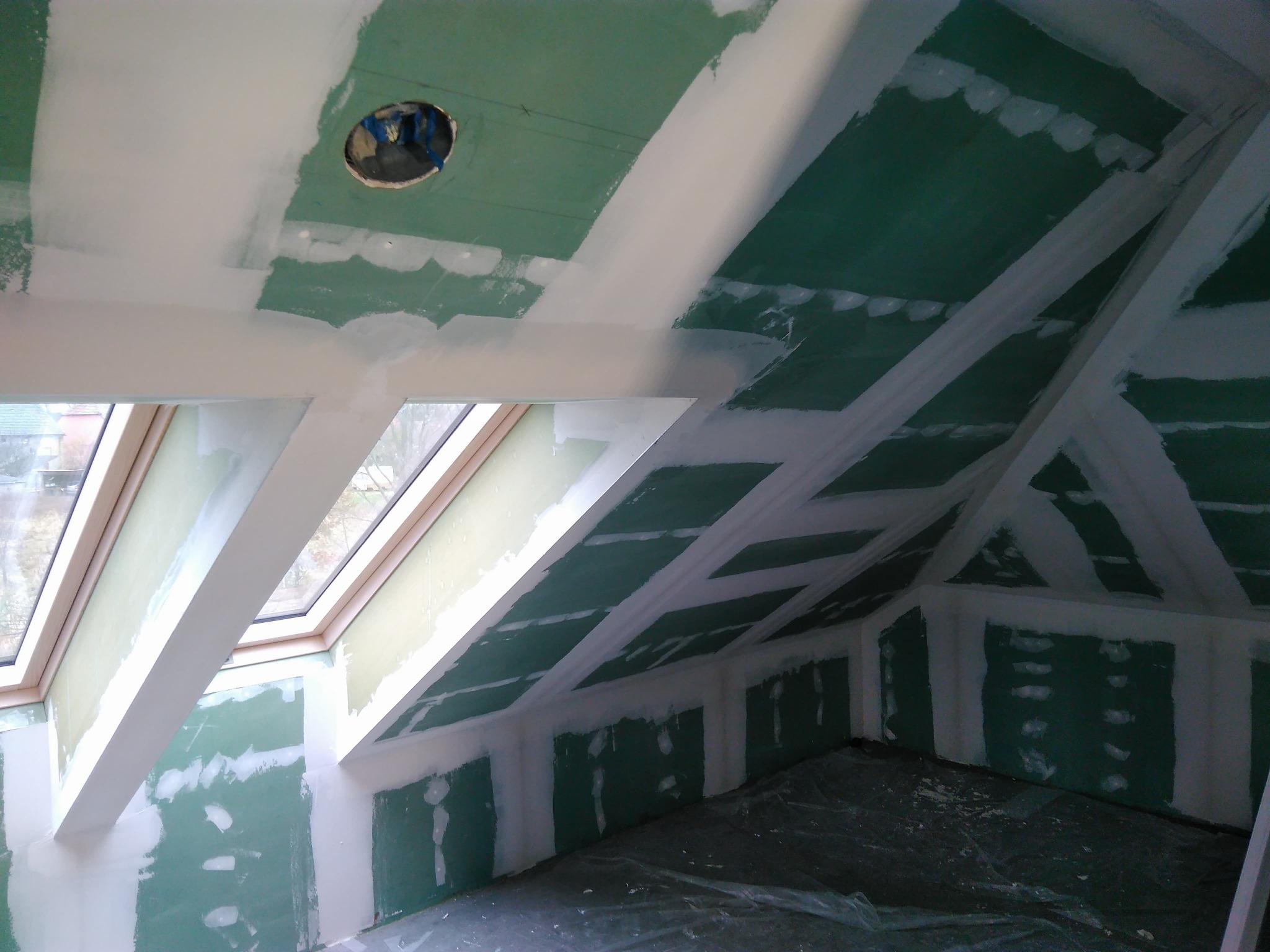 Poddasze w trakcie wykańczania: zielone płyty gipsowo-kartonowe na ścianach i suficie, częściowo pokryte białą szpachlą, widoczny otwór na oświetlenie w suficie i okno dachowe.