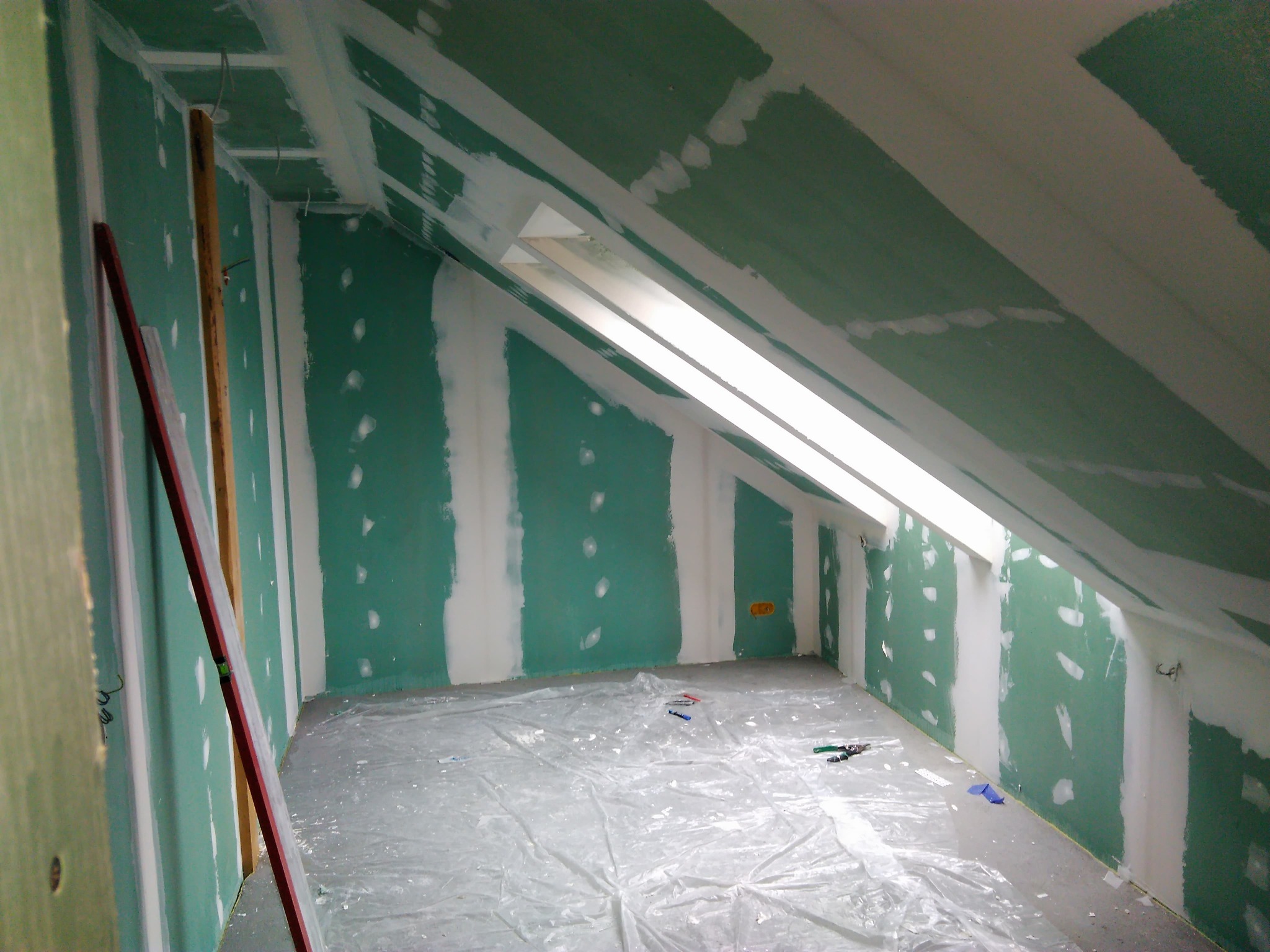 Pokój na poddaszu w trakcie remontu: zielone płyty gipsowo-kartonowe częściowo pokryte szpachlą, folia ochronna na podłodze, widoczne narzędzia i poziomica oparta o ścianę.