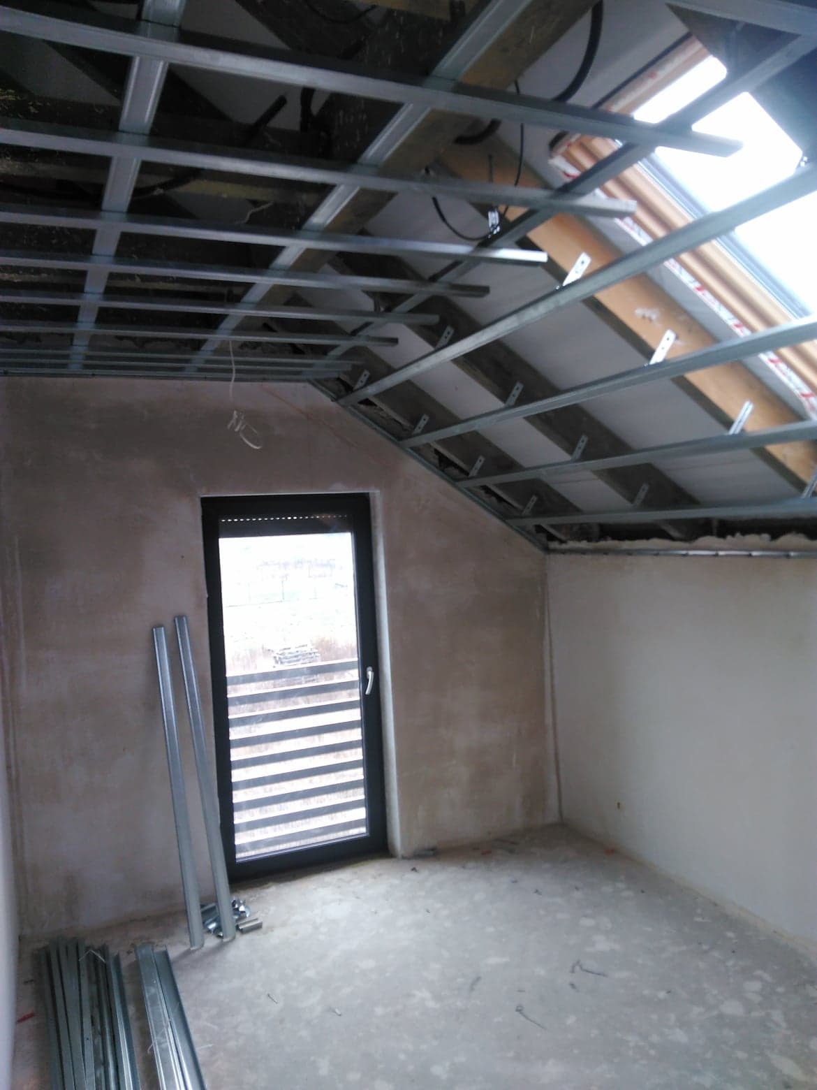 Poddasze w trakcie adaptacji: widoczne metalowe profile sufitowe podwieszane, okno dachowe, ściany w stanie surowym, drzwi z żaluzjami wewnętrznymi, profile metalowe na podłodze.