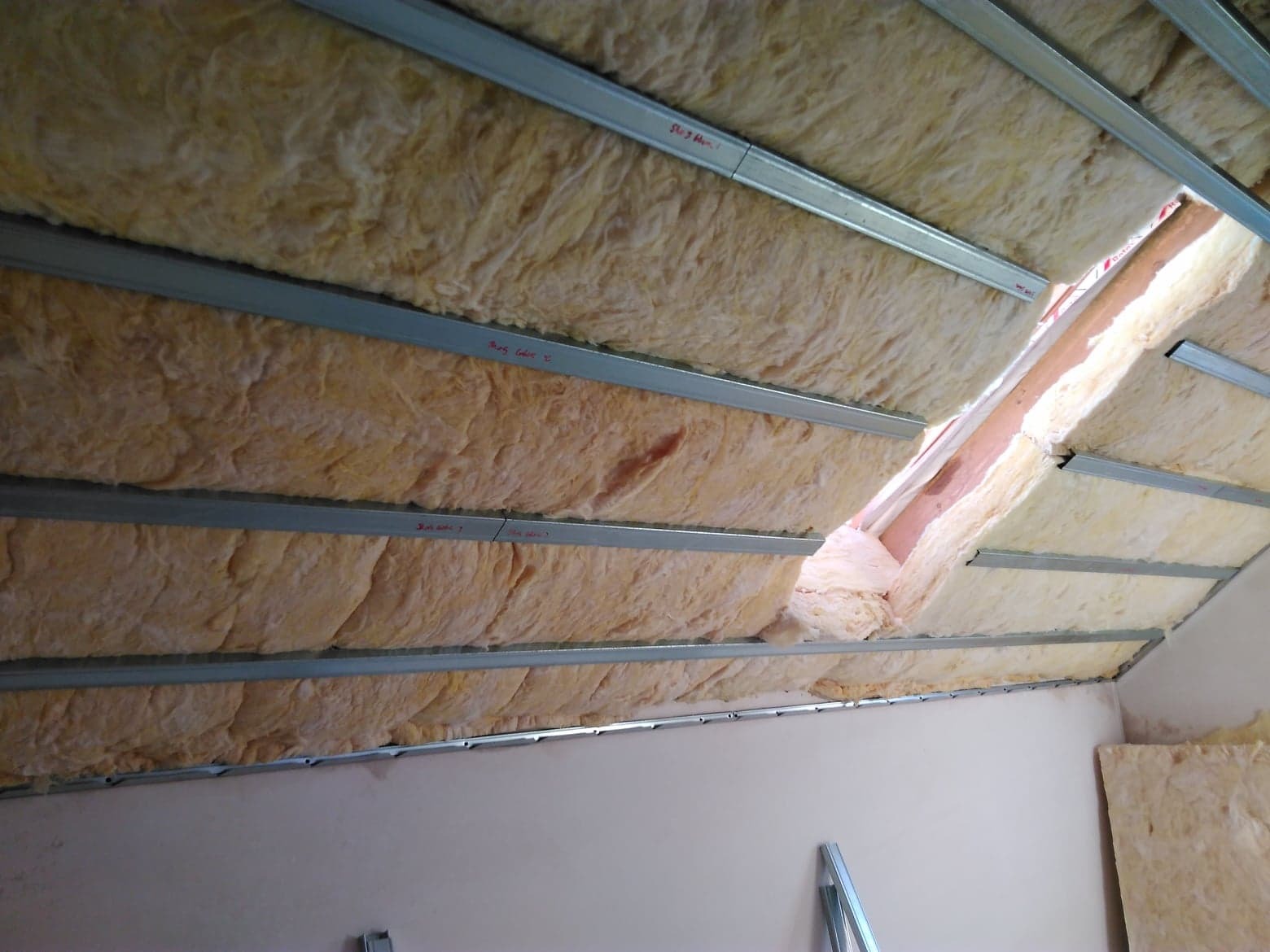 Izolacja poddasza wełną mineralną, widoczne metalowe profile montażowe i fragment ściany, etap prac wykończeniowych.