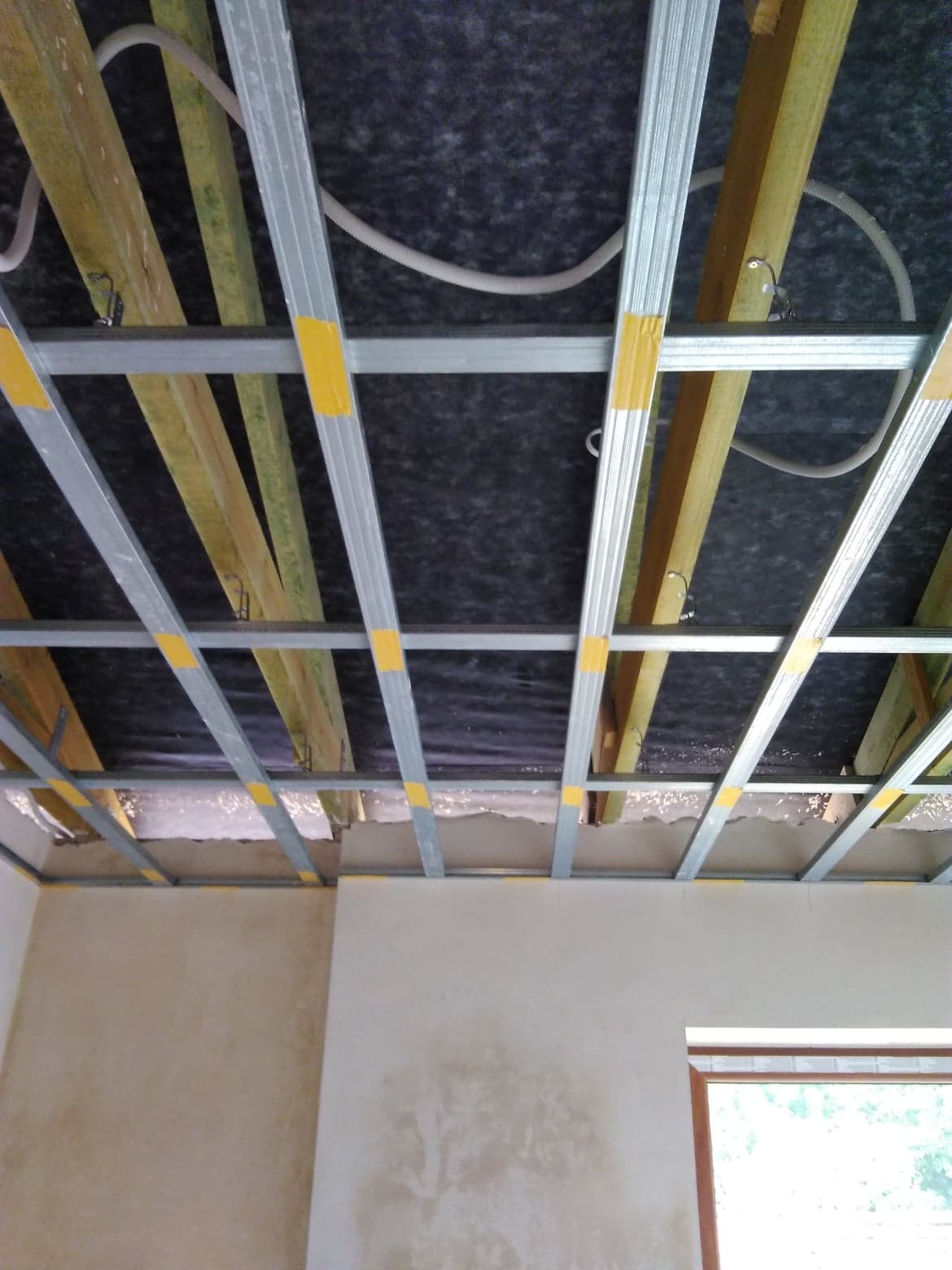 Konstrukcja sufitu podwieszanego z profili metalowych, zabezpieczonych żółtą taśmą, widoczne drewniane belki i instalacja elektryczna, przygotowanie do montażu płyt gipsowo-kartonowych.